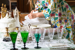 Table styling lessons in London. Woman in a floral robe, arranging items on a table with green glasses and a patterned wall in the background