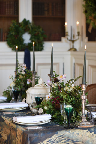 A styled holiday table setting with greenery, white flowers, tall black taper candles in silver holders, folded white napkins, and dark green glassware in an indoor room with a fireplace and wreath in the background.