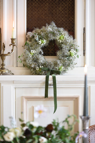 A decorative wreath made of pale green and white foliage with small white flowers and a dark green ribbon bow, hanging on a white fireplace mantel in an indoor setting with candles visible.