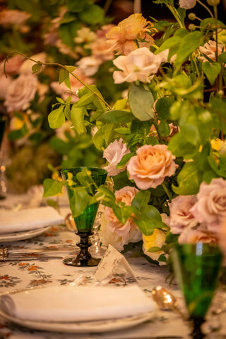 A close-up of an elegant floral centerpiece with blush pink roses and green leaves on a decorated table, accented with green glass goblets and white plates.