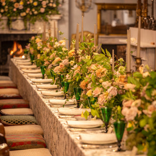 A long formal dining table set with white plates, green glass vases filled with pink and cream floral arrangements, and candles, photographed in an elegant indoor room with a fireplace and upholstered chairs visible along the left side.