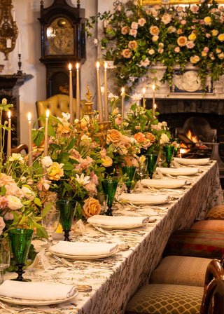 A long dining table set for guests in a warm, elegant room with a fireplace and floral arrangements. The table is covered with a patterned tablecloth, white plates, folded white napkins, green glass goblets, and multiple floral centerpieces in soft peach, pink, yellow, and green tones.