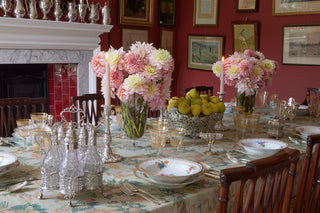 A styled dining table set for a formal dinner, featuring pink and cream floral bouquets, a bowl of pears, clear glassware, and floral plates on a patterned tablecloth in a dining room with a red wall and fireplace.