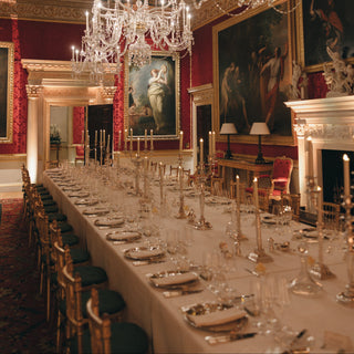An ornate dining room with a long table set for many guests under large crystal chandeliers, red walls, classical paintings, and gilded details.