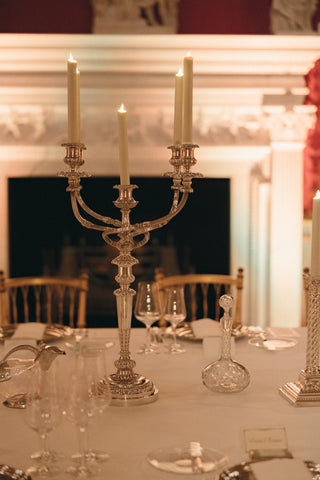 An ornate silver candelabra holding lit taper candles sits on a white tablecloth set dining table in a warmly lit room with a fireplace and gold chairs.
