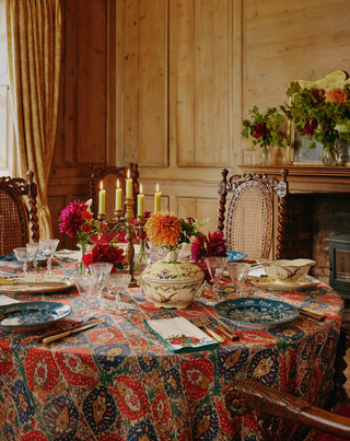 A round dining table set for a meal in a rustic wood-paneled room. The table is covered with a colorful paisley-patterned tablecloth and surrounded by ornate carved wooden chairs, floral centerpieces with red and orange flowers, lit candles, and decorative dishes.