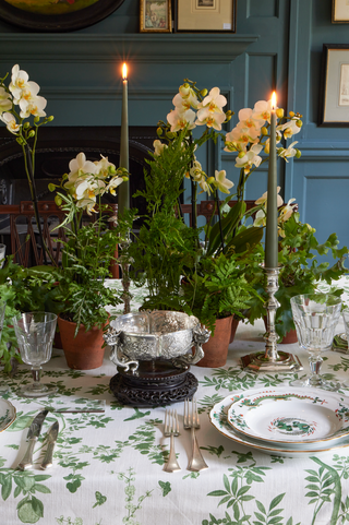 A styled dining table setting with a green and white botanical tablecloth, silver cutlery, crystal glasses, stacked plates, potted green plants, and tall green taper candles in silver holders. A silver bowl on a dark carved base sits in the center, with white orchids rising above the arrangement against a teal paneled wall background.