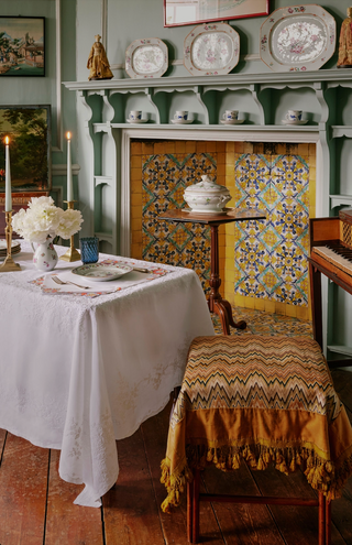 A styled interior scene with a white tablecloth-covered table, a patterned yellow and brown fringed textile draped over a wooden chair, a fireplace with decorative tiles and wall plates, and a piano edge visible on the right.